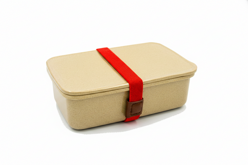 A rectangular beige bento-style lunch box with a matching lid, secured by a bright red elastic strap with a small brown buckle, shown on a plain white background.