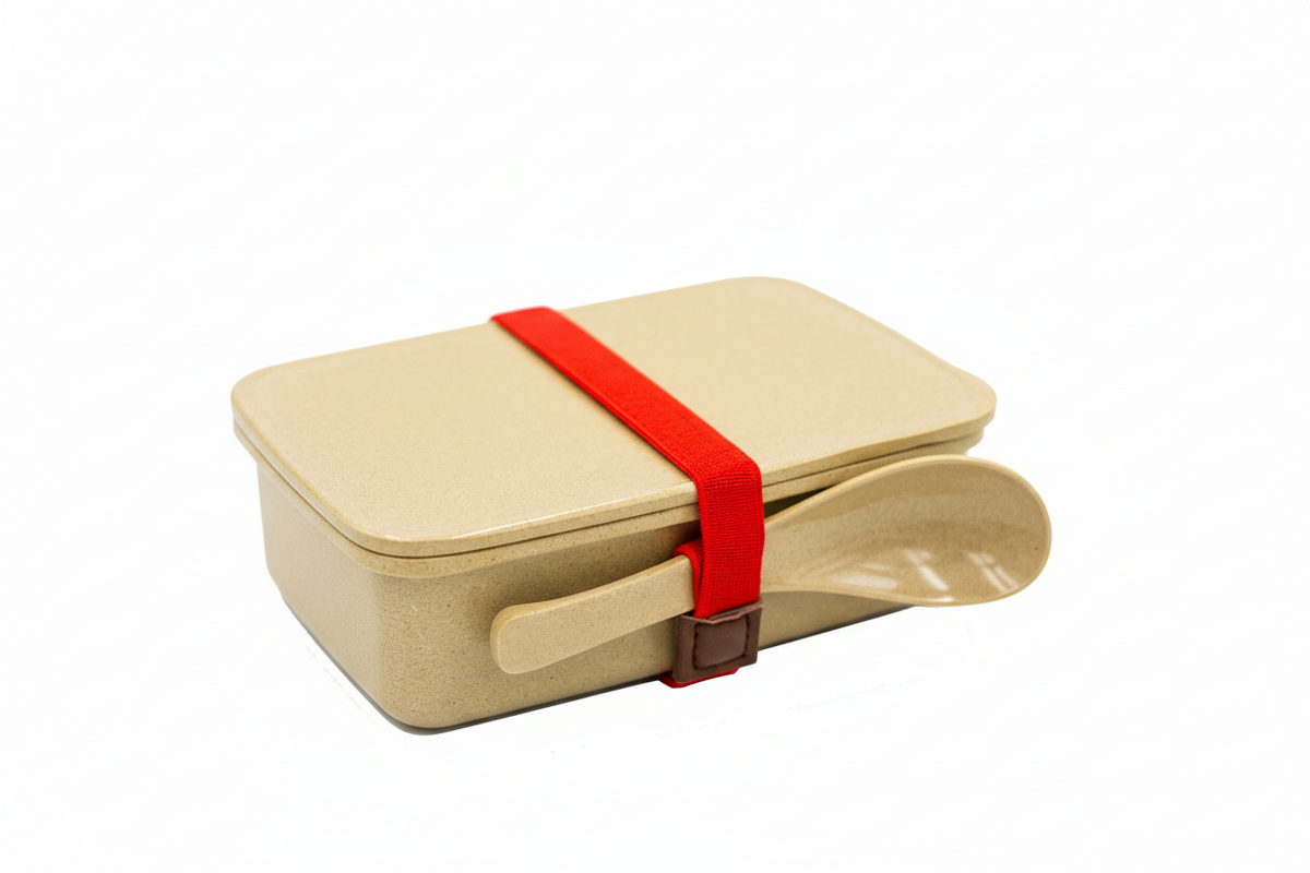 A light beige bamboo-style lunch box with a matching lid, secured by a bright red elastic band, and a matching beige spoon resting in front, shown on a plain white background.