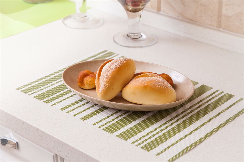 A green and white striped placemat on a white countertop, holding a beige plate with three golden-brown bread rolls and two small pastries. A wine glass base is blurred in the background.