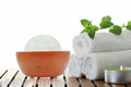A wooden diffuser with a frosted glass top sits on a slatted wooden surface beside rolled white towels, mint leaves, and a small lit tealight candle against a white background.