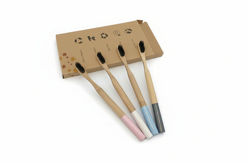 Four bamboo toothbrushes with colorful handles in pink, white, light blue, and dark gray, arranged in front of a brown cardboard box on a white background.