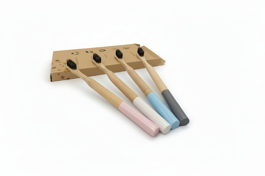 Four bamboo toothbrushes with pastel-colored handles in pink, white, light blue, and dark gray, arranged in front of a small cardboard box on a light gray background.