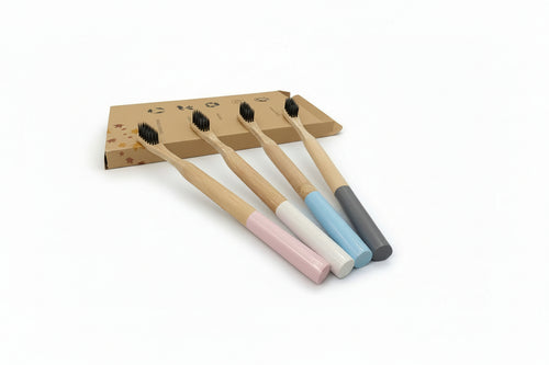 Four bamboo toothbrushes with pastel-colored handles in pink, white, light blue, and dark gray, arranged in front of a small cardboard box on a light gray background.