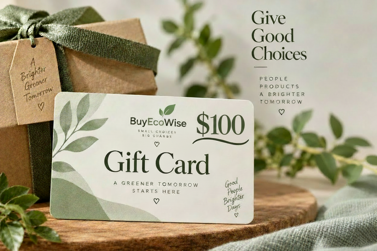 A BuyEcoWise $100 gift card displayed on a wooden surface next to a brown kraft gift box tied with a green ribbon. The scene includes soft-focus greenery and text on the right reading 'Give Good Choices People Products A Brighter Tomorrow.